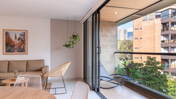 A modern living room with a sofa, a dining table, and two chairs. There is a large window with a view of a cityscape and a balcony with a hanging plant.