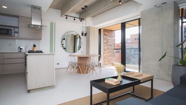 A modern kitchen with a microwave, stove, and wooden cabinets. There is a dining area with a round mirror, a table, and chairs. The room has large windows and a view of the city. - Amazing w Balcony 3 BR in Laureles Medell n (Medellín)