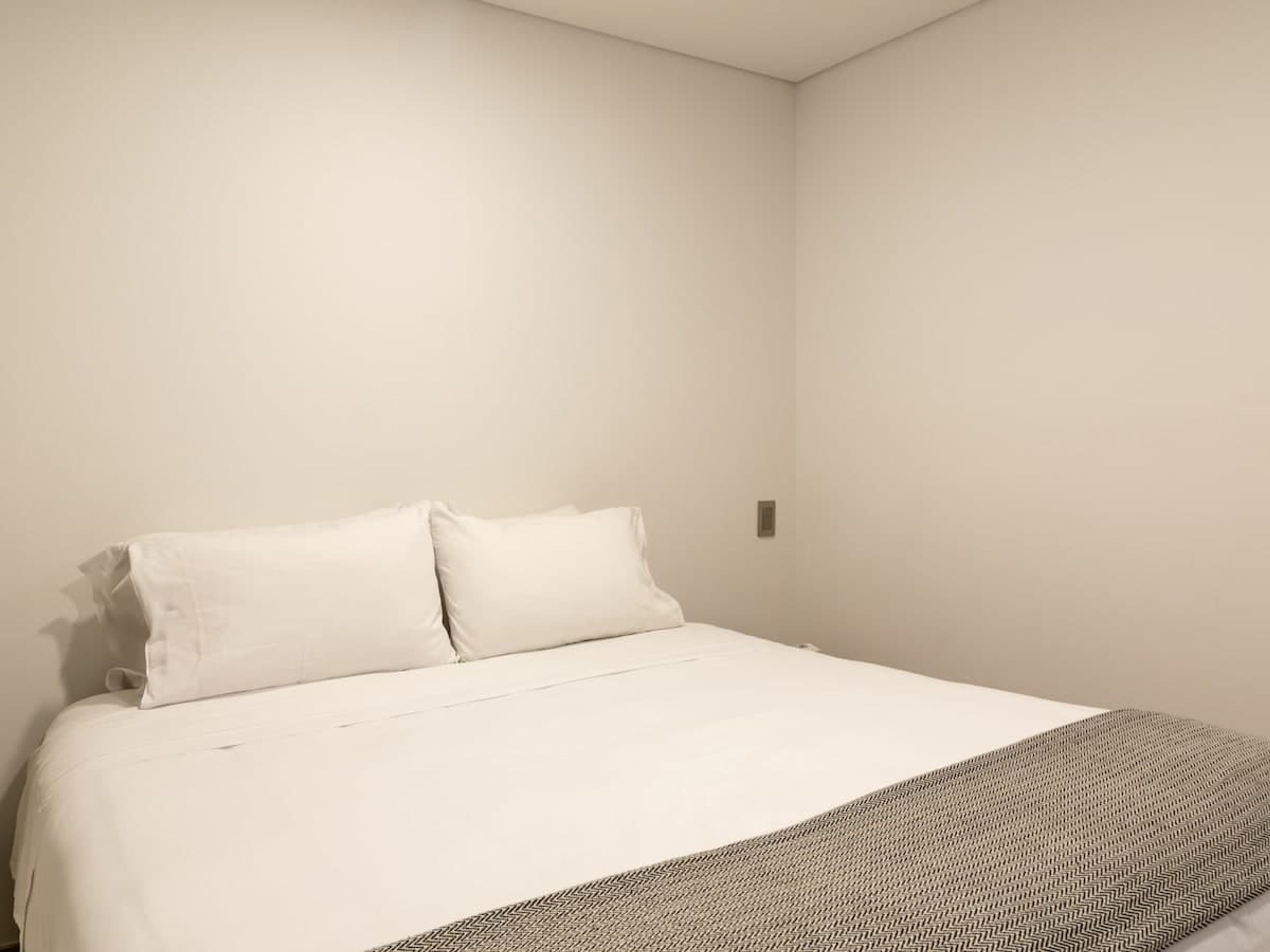 A simple, unadorned bedroom with a large bed, two pillows, and a plain wall.