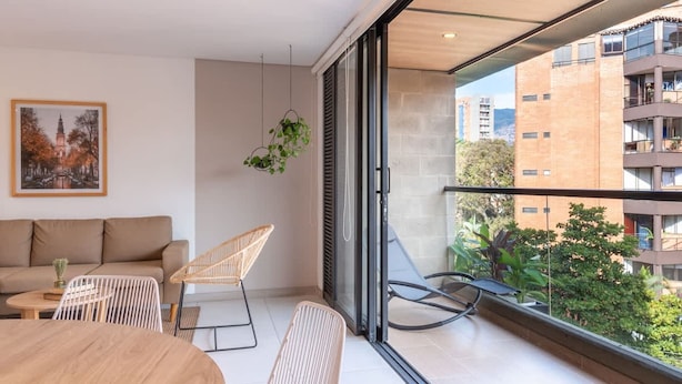 A modern living room with a sofa, chairs, a dining table, and a large window with a city view. - Bright 3BR in Laureles Medell n (Medellín)