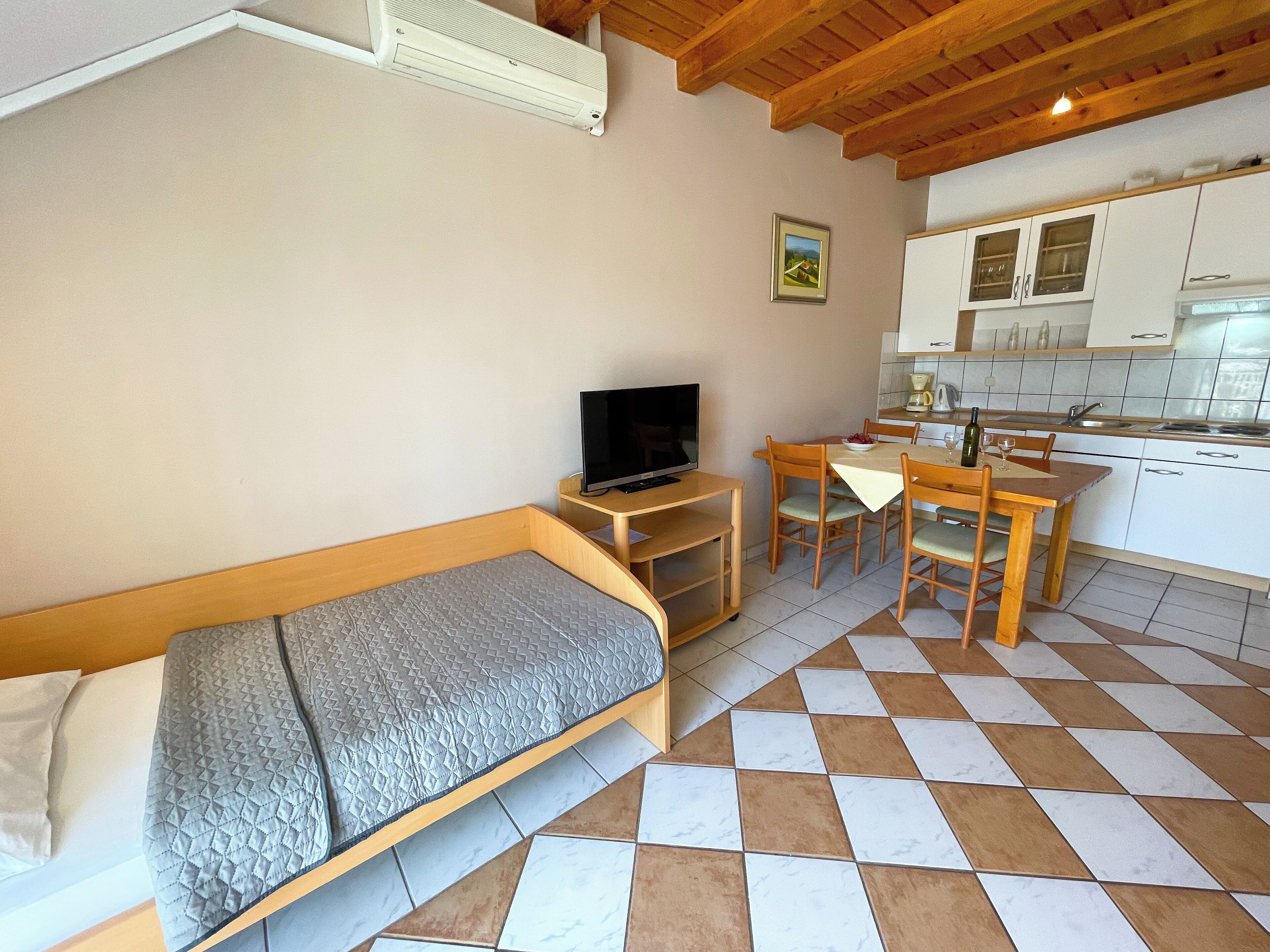 A compact room with a bed, a TV, a dining table with chairs, and a kitchenette.