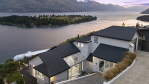 Ett modernt hus med ett stort glasfönster, ett svart tak och utsikt över en sjö och berg.