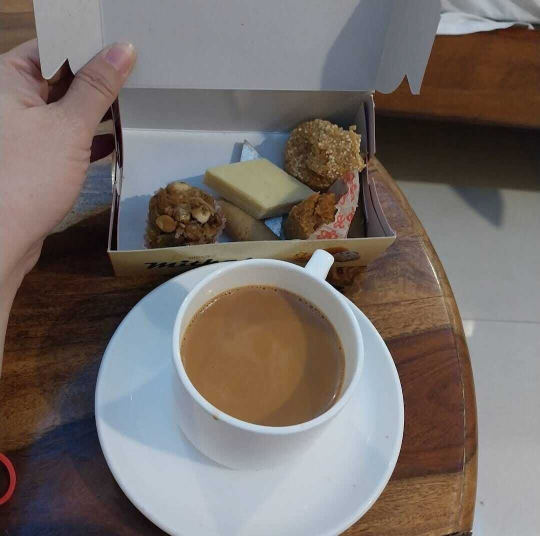 A hand holding a box of food items, including fried snacks and a bar of soap, next to a cup of coffee on a wooden table.