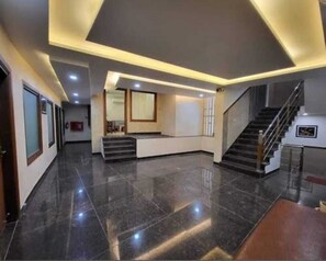 A spacious lobby with a polished floor, a central staircase, and a reception desk.