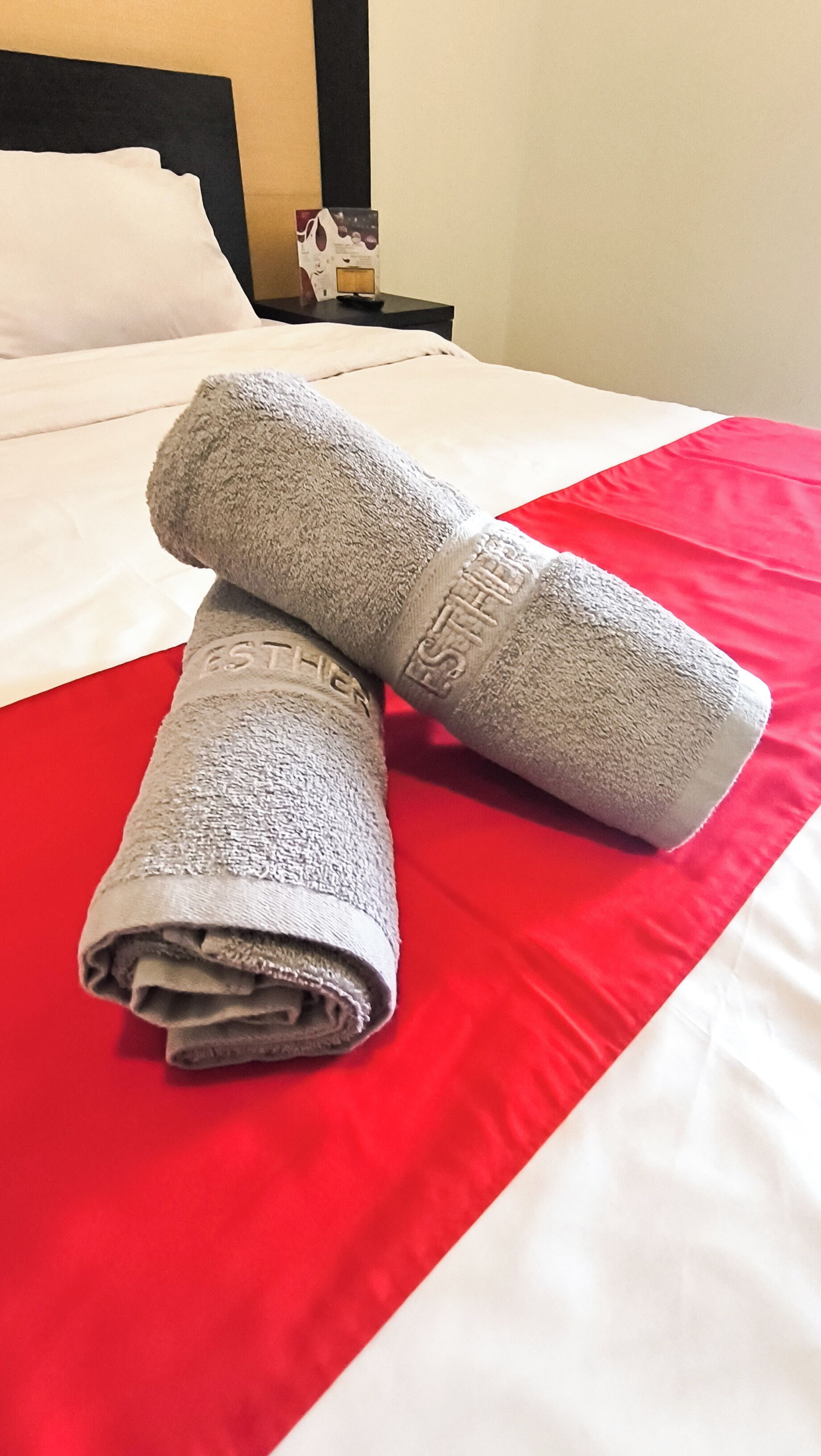 A neatly made bed with a red and white bedspread, two rolled towels with the word "ESTHETICS" embossed on them, and a small nightstand with a figurine in the background.