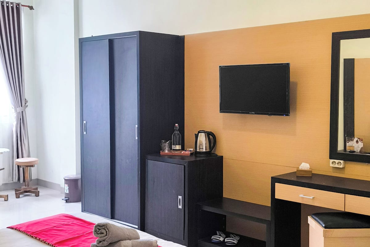 A modern hotel room with a large bed, a flat-screen TV mounted on the wall, a wardrobe, a desk with a mirror, and a red accent runner on the bed.