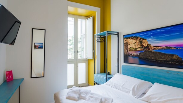 A hotel room with a bed, a desk, a TV, a window, and a painting of a coastal scene. - Central Station Housing Apartments (NAPOLI)