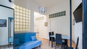 A modern living room with a blue sofa, a dining table with chairs, a chandelier, and a large window with blinds. - Central Station Housing Apartments (NAPOLI)