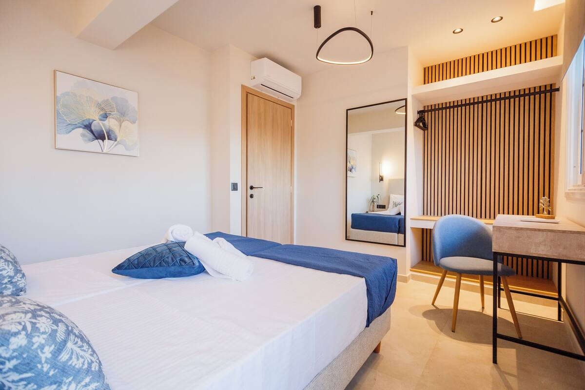 A hotel room with a bed, a desk, a chair, a mirror, and a wall-mounted air conditioner.