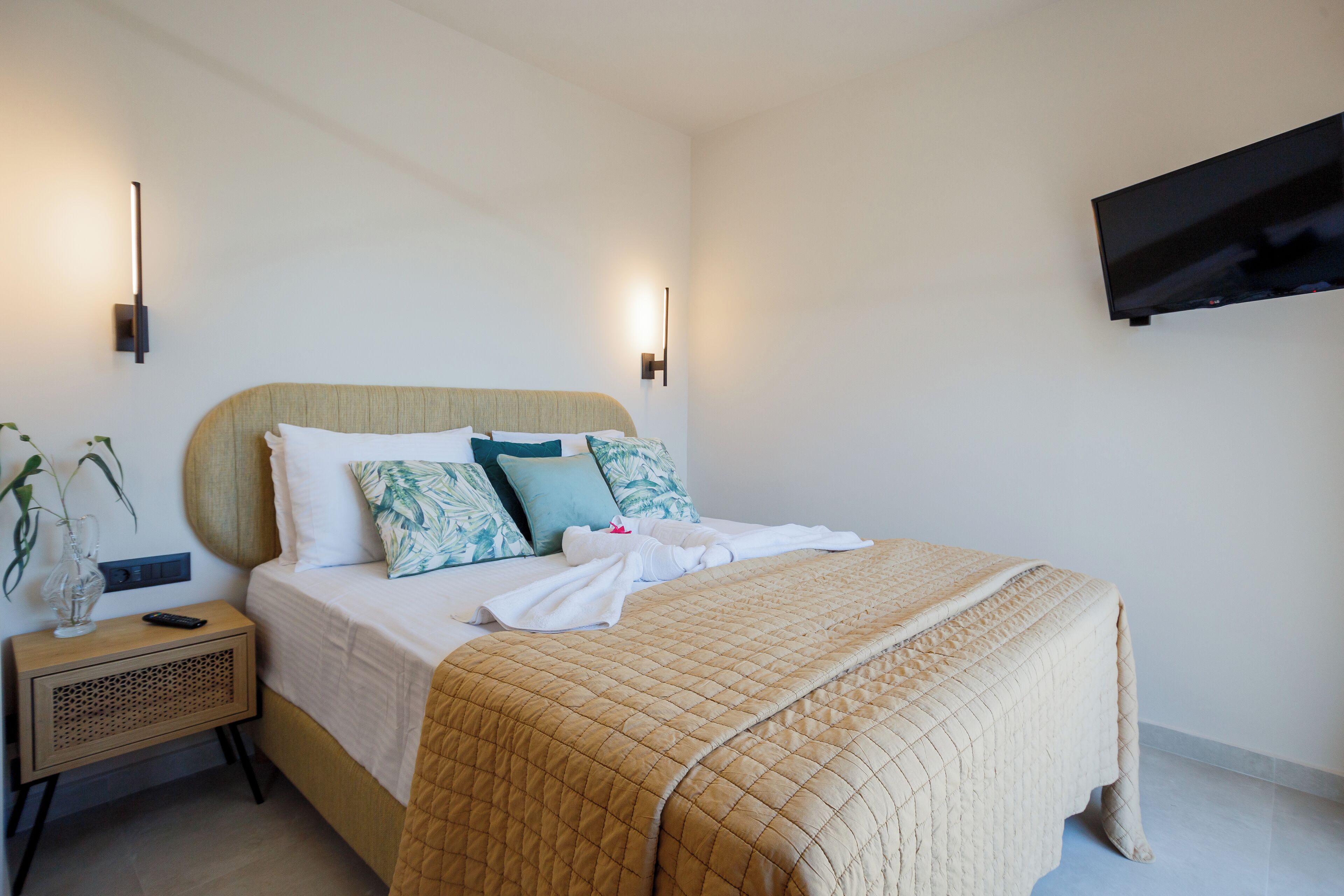 A neatly made bed with a quilted blanket, a bedside table with a vase and remote control, and a wall-mounted TV.