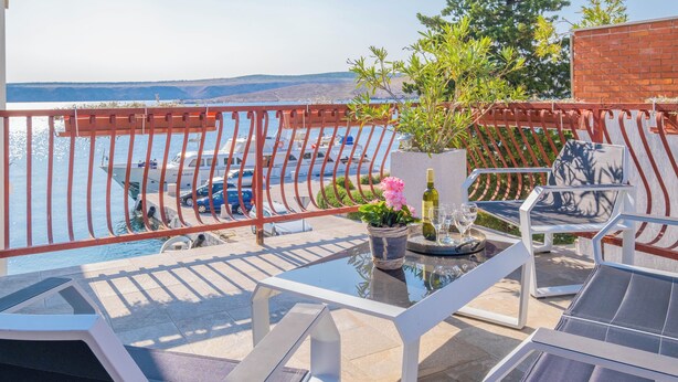 Un balcón con una mesa de vidrio, sillas, una planta en maceta y vistas al mar y a las montañas.