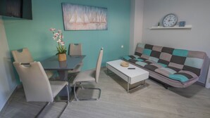 Una sala de estar con paredes color turquesa, un sofá blanco, una mesa de comedor con sillas, un televisor y un cuadro con barcos de vela.