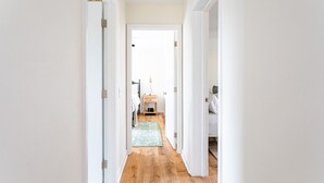 Um corredor com piso de madeira que leva a um quarto com cama, mesa de cabeceira e criado-mudo.
