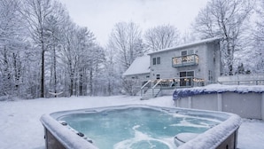 Quintal coberto de neve com hidromassagem e uma casa com varanda.