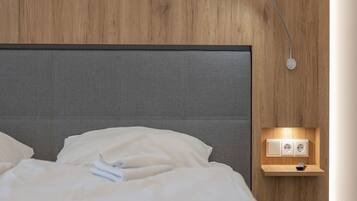 A neatly made bed with white linens and a gray headboard. There is a wall-mounted nightstand with a lamp and a power outlet.