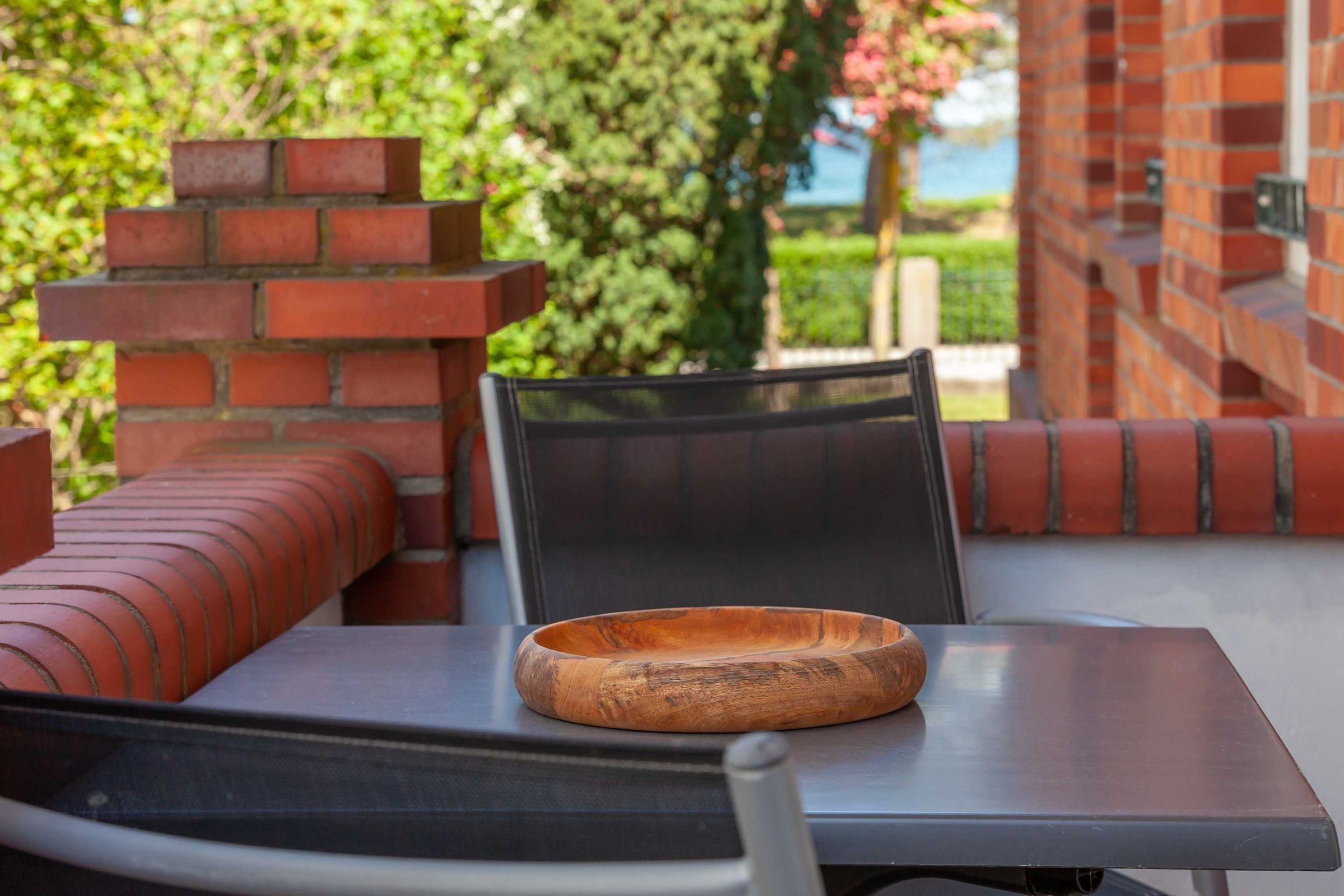 Eine Terrasse mit einer Holtschale auf einem Tisch, Ziegelmauern und Blick auf Grün.