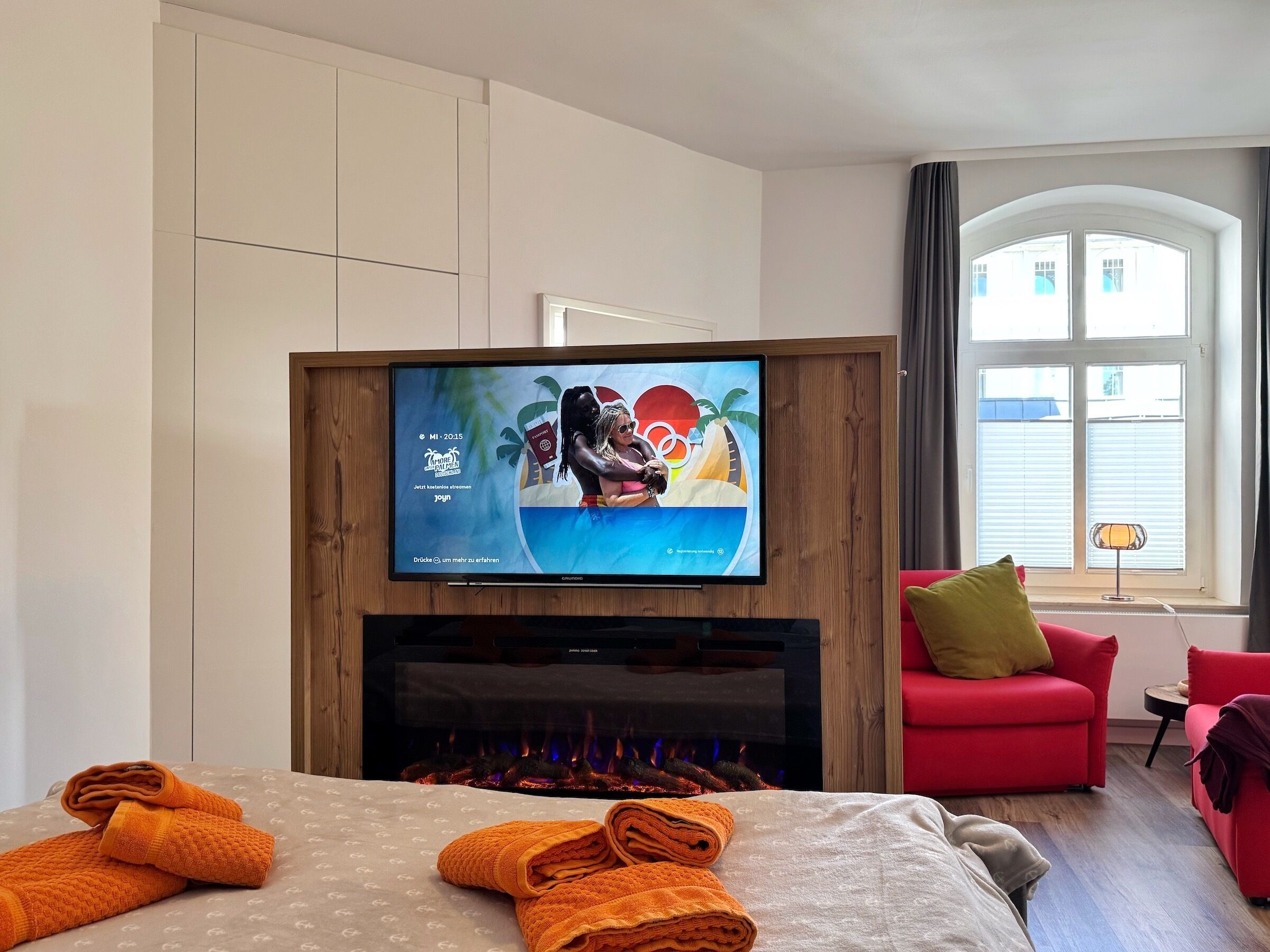 Habitación de hotel con televisión incorporada en un panel de madera, chimenea, cama con toallas anaranjadas, sillón rojo y vistas al exterior a través de amplias ventanas.