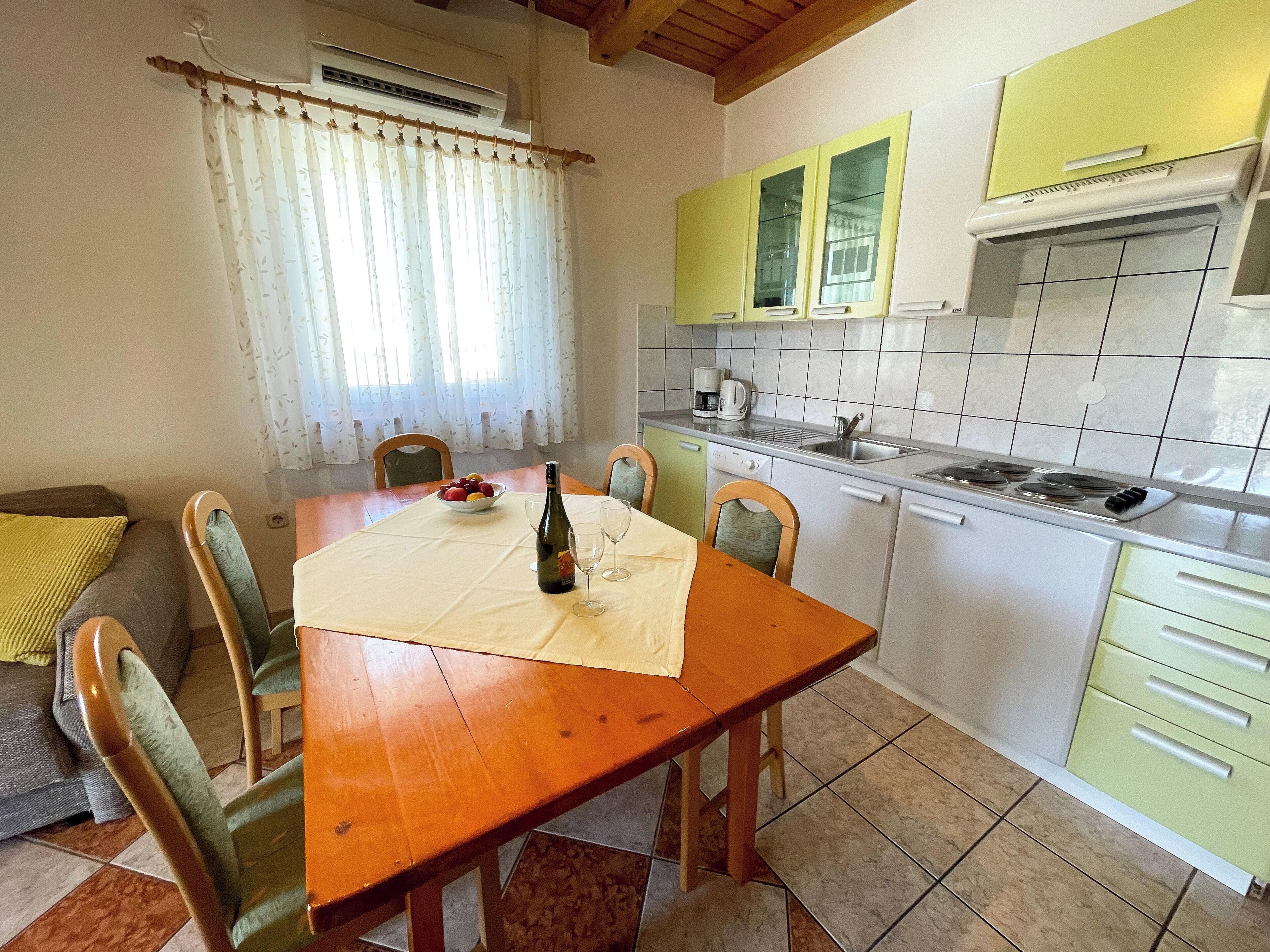 A dining area with a wooden table, chairs, a bowl of fruit, a bottle of wine, and a coffee maker.