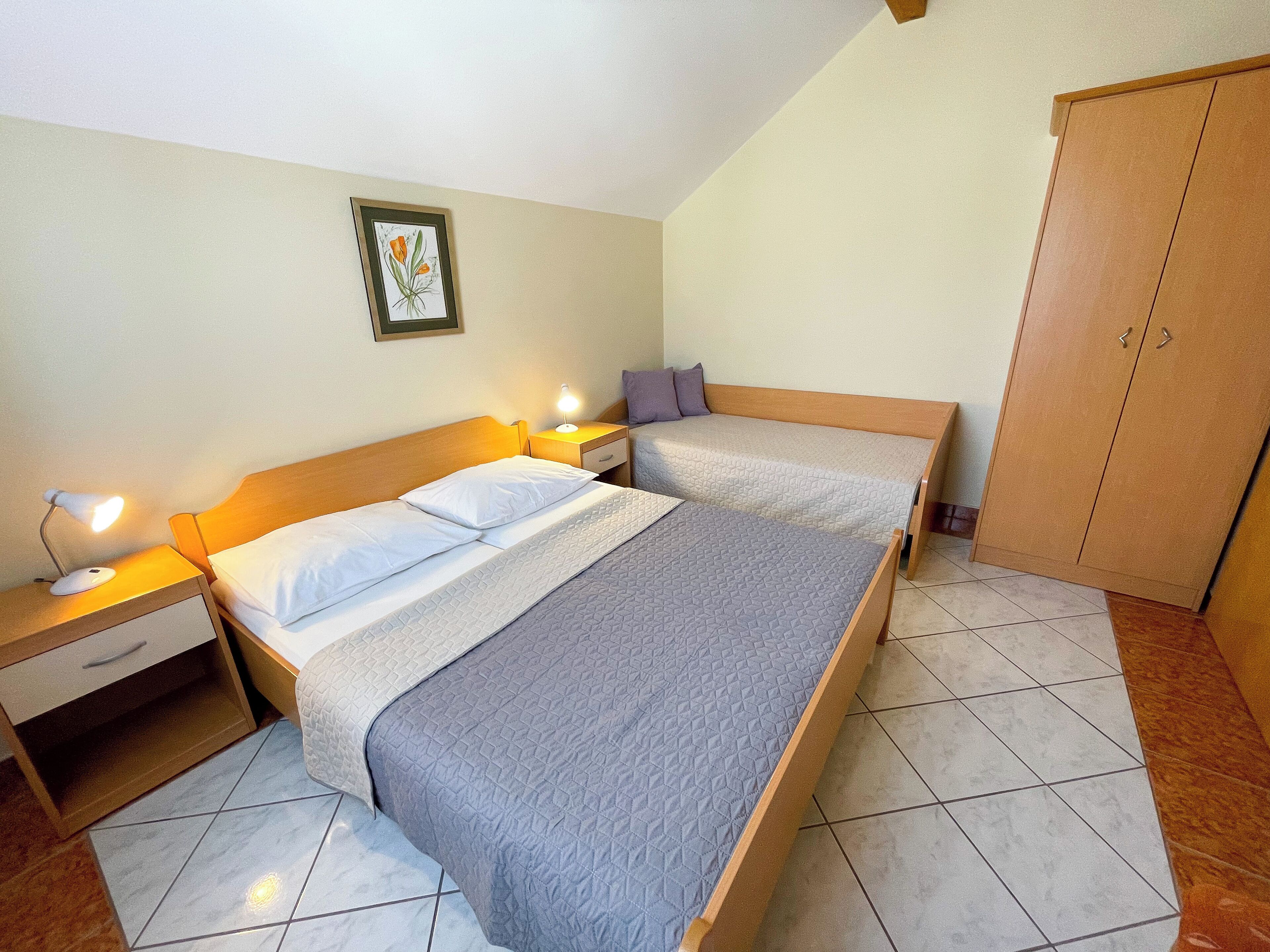 Une chambre avec deux lits, une table de chevet avec une lampe, une armoire et une estampe botanique encadrée au mur.