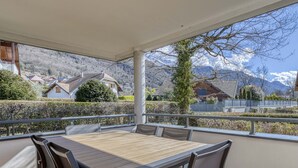 Une terrasse extérieure couverte, avec une table et des chaises en bois, donnant sur un quartier résidentiel avec des maisons et des montagnes.