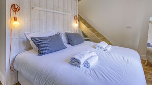 Une chambre à coucher avec une tête de lit en bois, du linge de lit blanc, des coussins à carreaux, une serviette enroulée et un escalier en bois.