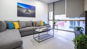 Un salon moderne avec un canapé gris, des coussins colorés, une table basse et une grande fenêtre donnant sur des immeubles.