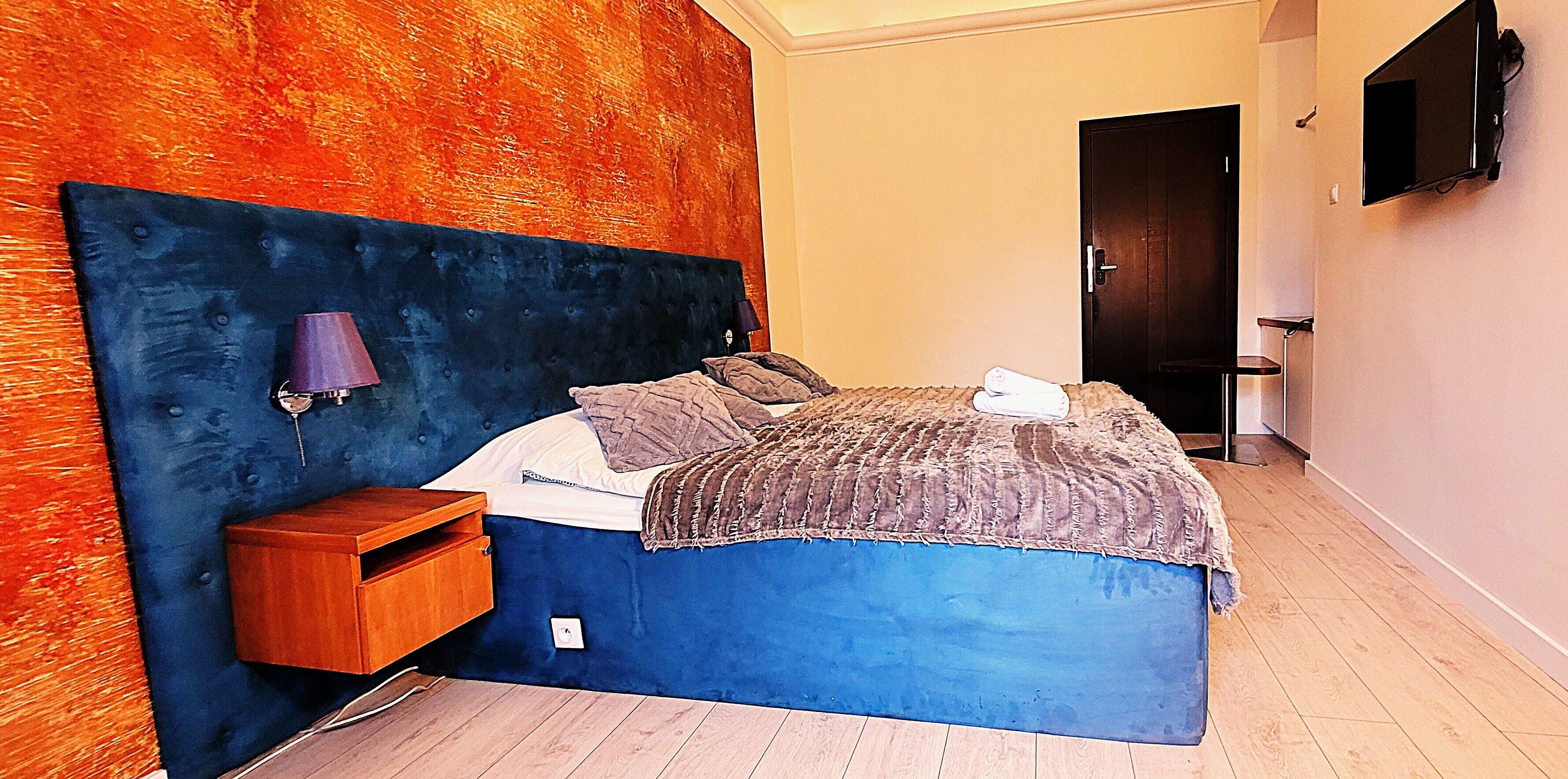 Une chambre avec une tête de lit bleue, une table de chevet en bois, un lit recouvert d’une couverture blanche et marron, et un téléviseur fixé au mur.