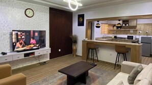 A modern living room with a flat-screen TV, a dark coffee table, and a sofa. There is a kitchen area with a bar and stools. - Me Time Residence (Enugu)
