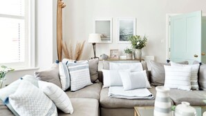 Ein gemütliches Wohnzimmer mit einem beigefarbenen Sofa, weißen Kissen, einem Beistelltisch mit Lampe, gerahmten Bildern an der Wand und einer Tür mit hellblauer Umrandung.