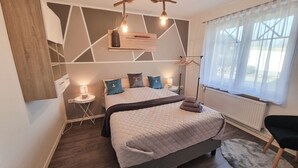 Ein Schlafzimmer mit Bett, Nachttischlampen, einer Pendelleuchte, einem Heizkörper und einem Fenster mit transparenten Vorhängen.