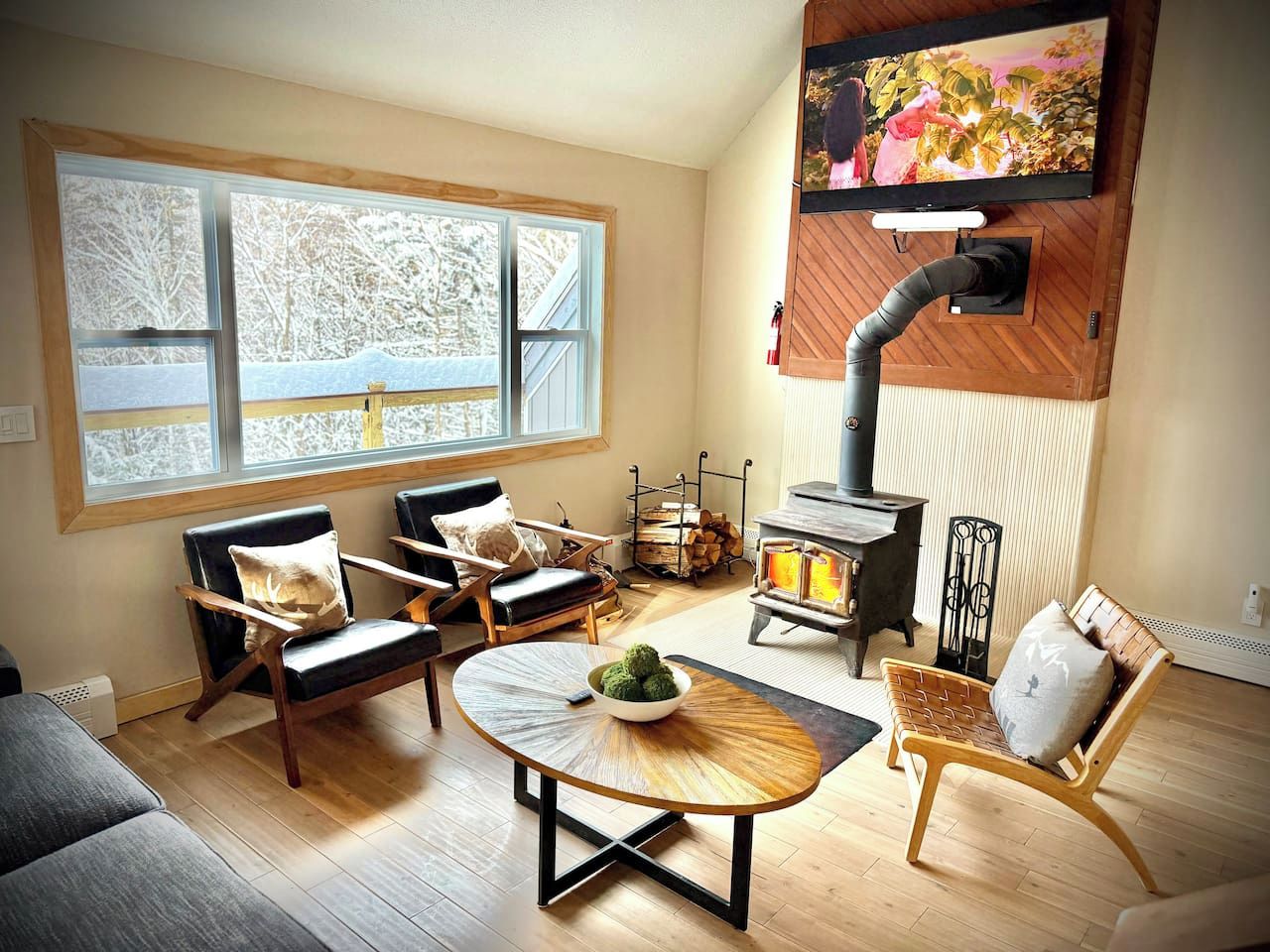 Un salon chaleureux avec une cheminée, des meubles en bois et une grande fenêtre donnant sur des arbres couverts de neige.