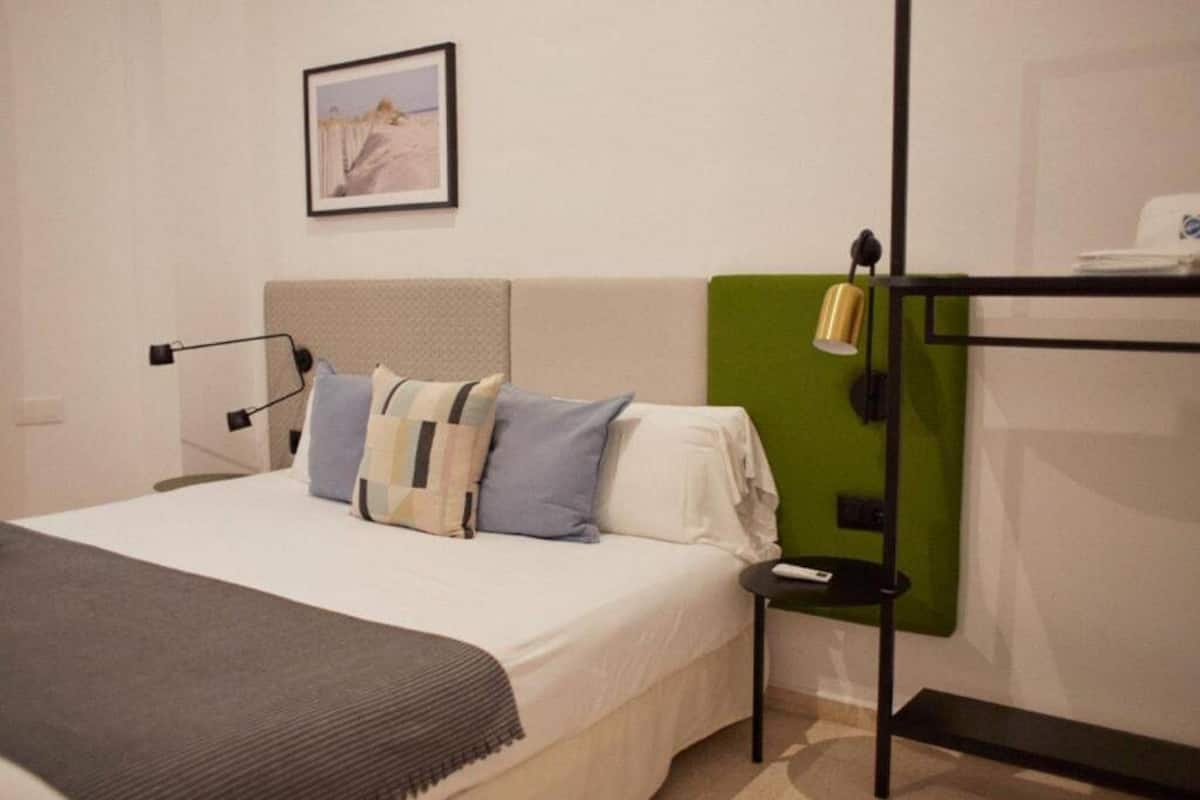 Une chambre d’hôtel bien aménagée, avec un lit, une table de chevet, un panneau mural vert, un tableau encadré au mur et une petite table avec une lampe.