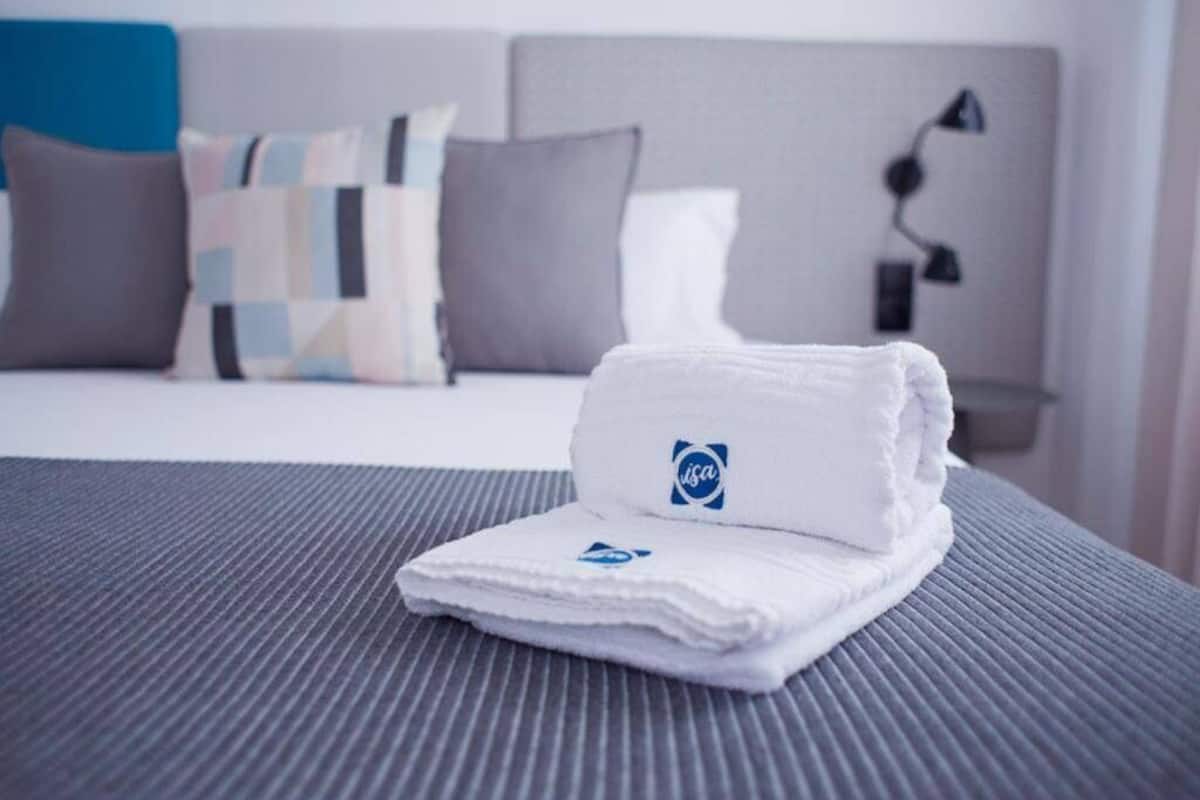 Un lit bien fait, avec une serviette blanche pliée et un logo dessus, une couverture à rayures grises et, à l’arrière-plan, un lit avec une tête de lit grise et des oreillers.