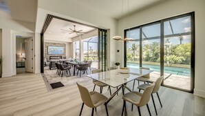 A spacious dining area with a large table and chairs, a kitchen in the background, and a view of a swimming pool through large windows. - Villa Sunrays -NEW 3-bedroom Villa with pool & spa (Cape Coral)