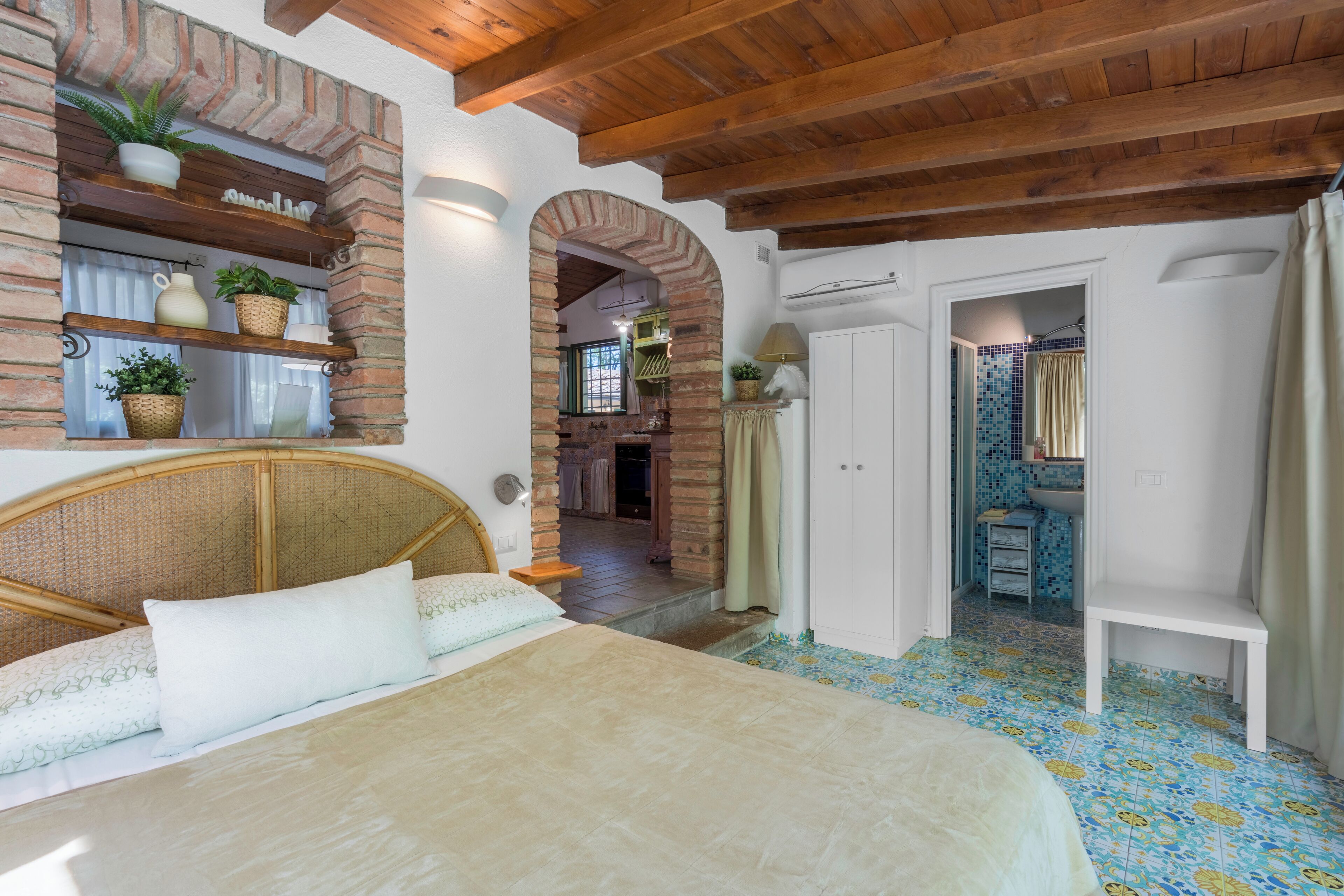 A bedroom with a bed, wooden headboard, brick shelving with plants, a bathroom with a tiled floor, and a wooden ceiling.