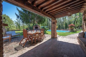 Una terrazza esterna con tavolo e sedie in mattoni, un camino in pietra e una piscina sullo sfondo.