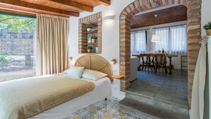 A bedroom with a bed, wooden ceiling beams, a brick archway, a dining area with a table and chairs, and a view of greenery outside. - Grace Home Pool Wifi Gambassi Terme (Gambassi Terme)