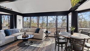 A modern living room with a sectional sofa, a round coffee table, a dining table with chairs, and large windows offering a view of trees and a building.