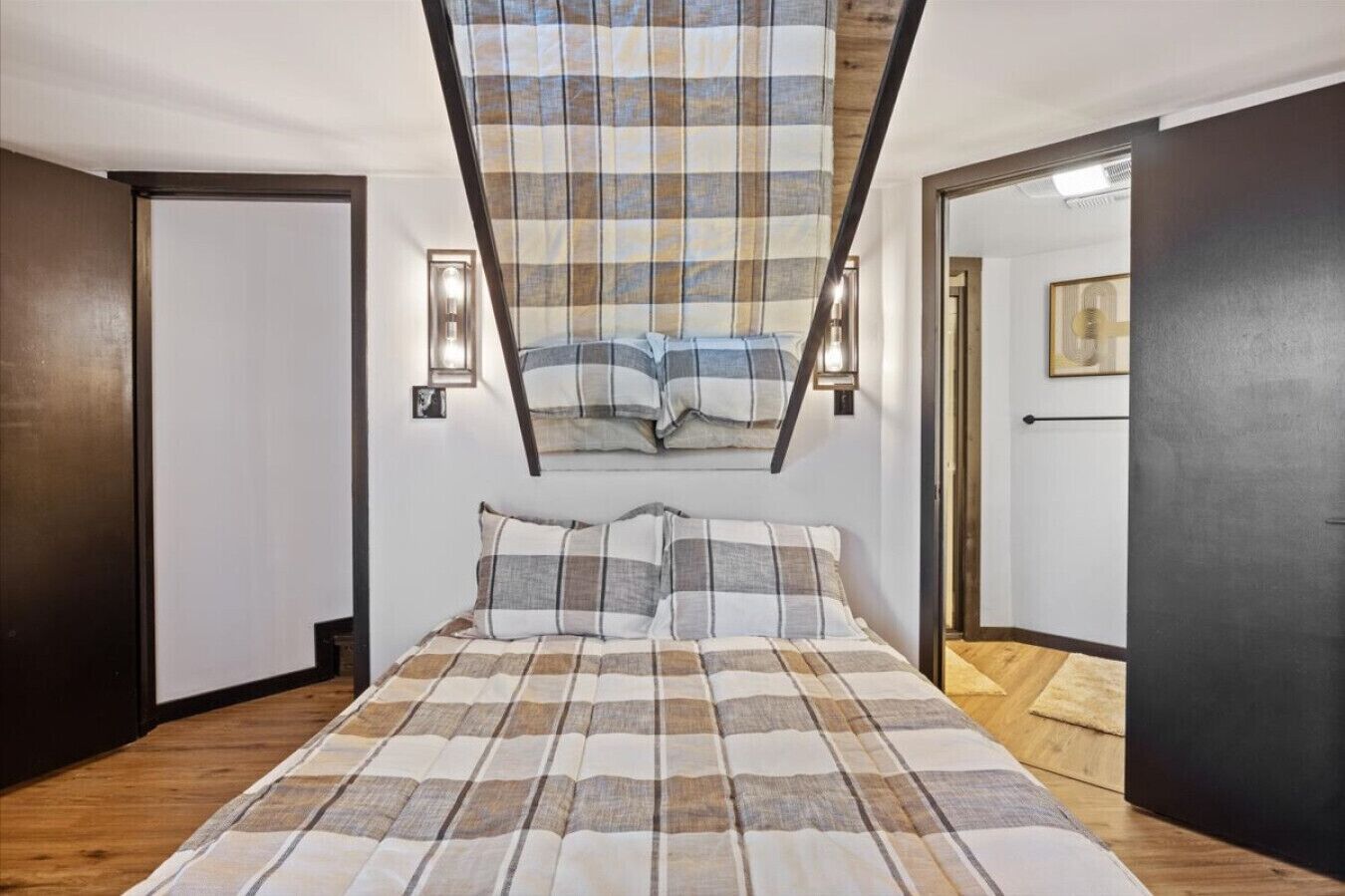 A bedroom with a bed, two wall-mounted lights, a window with a plaid curtain, and a door on the right.