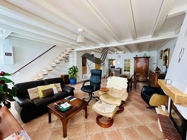A spacious living area with a black leather sofa, a wooden coffee table, a beige armchair, and a dining table. A staircase with white railings leads to an upper level. - Le Ferney - Close to downtown and the spa (Rochefort)