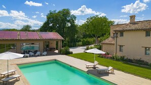 Una piscina con sillas de descanso y sombrillas, rodeada por un césped cuidado y un edificio con tejado de baldosas.
