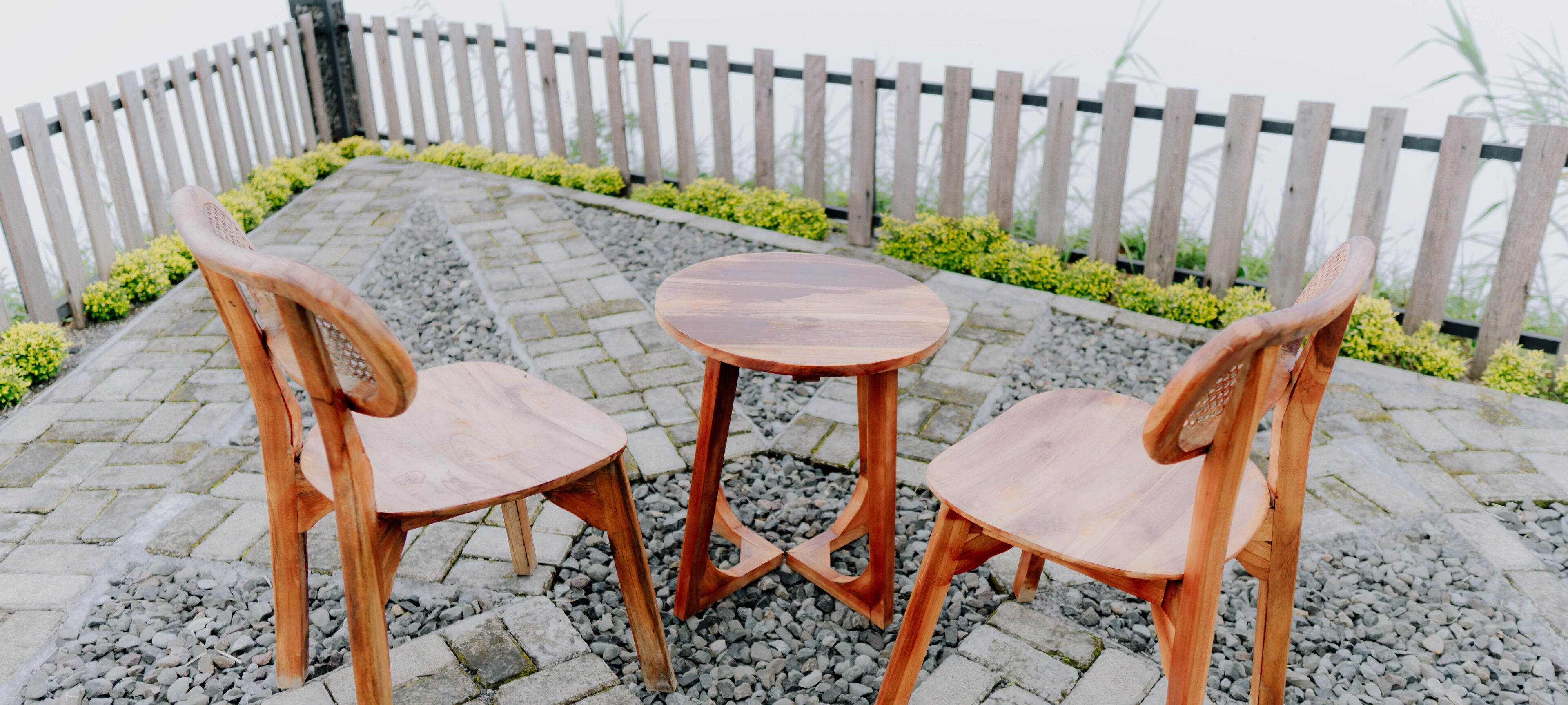 A wooden table and two chairs set on a stone patio with a wooden fence in the background.
