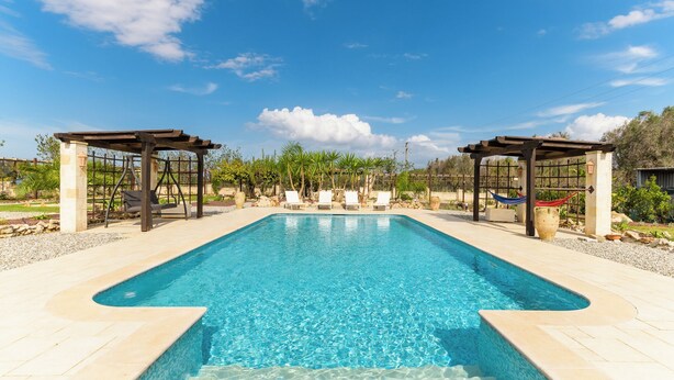 Une piscine rectangulaire à l’eau d’un bleu limpide, entourée d’une terrasse carrelée. On y trouve deux pergolas en bois avec des coins salon et un hamac.