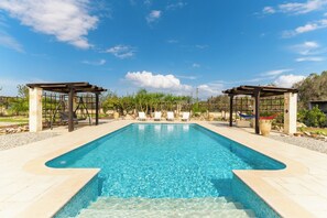 Een rechthoekig zwembad met helderblauw water, omgeven door een betegeld zonnedek. Er zijn twee houten pergola’s met zitgedeeltes en een hangmat.