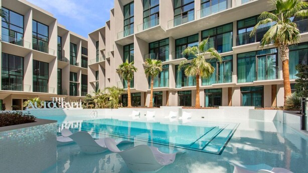 A modern building with multiple floors, a swimming pool with lounge chairs, and palm trees. - Chic Studio with Pool Access in The Autograph (Dubai)