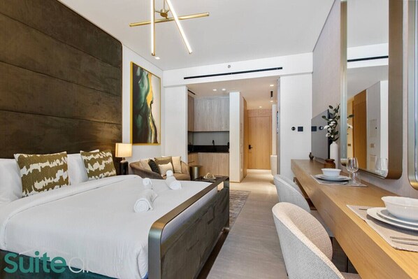 Quarto de hotel moderno com uma cama grande, área de estar com sofá, escrivaninha de madeira com espelho e vista para a cozinha.