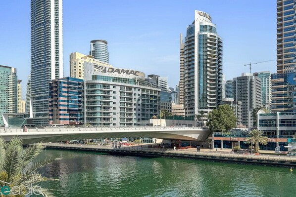 A modern cityscape with high-rise buildings, including a prominent building with 'DAMAC' signage. A pedestrian bridge spans over a water body, and there are palm trees visible.