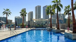 A swimming pool with a cityscape in the background, palm trees, and a clear sky.