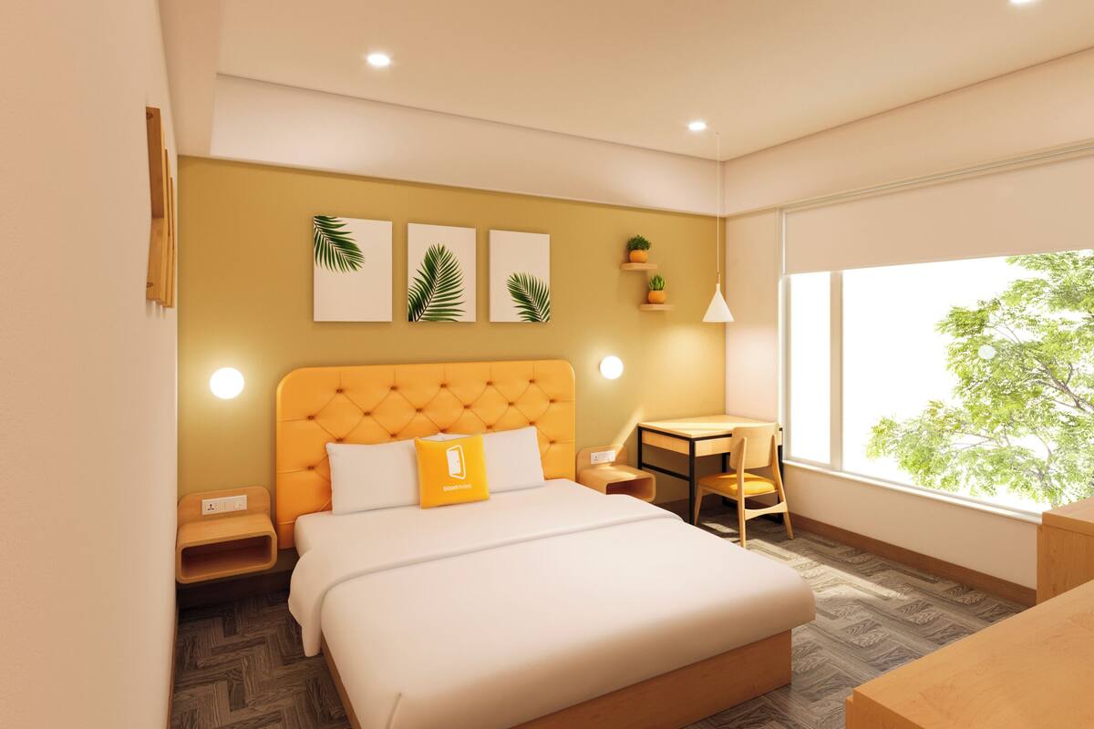 A modern hotel room with a large bed, a small table, and a chair. There are wall-mounted lights, a window with a view of trees, and a wall with framed pictures.
