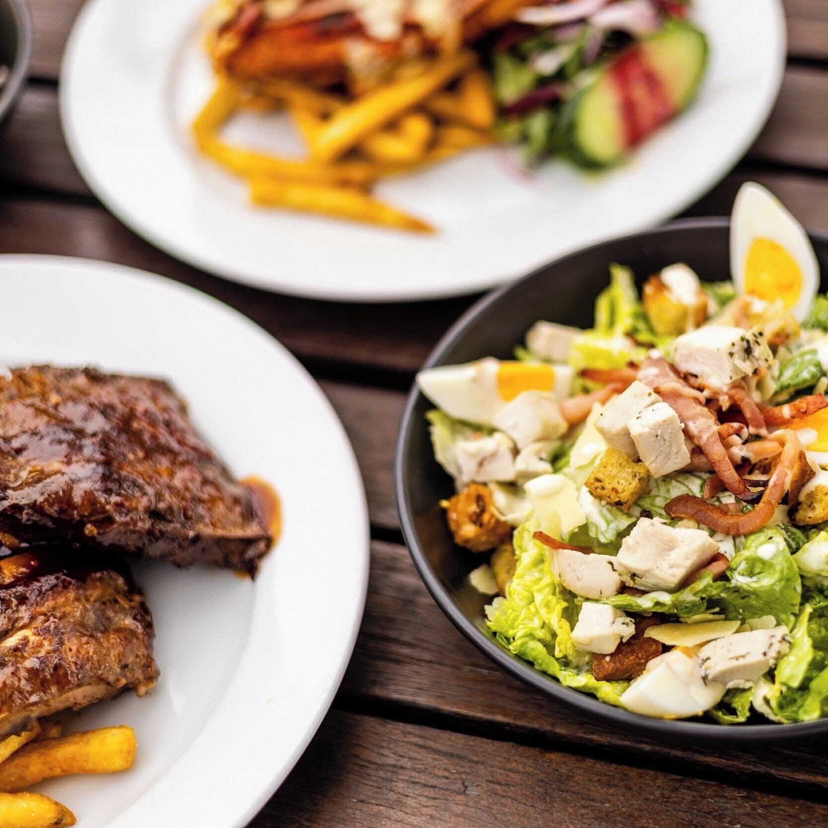 Una mesa con tres platos de comida: un plato con carne a la parrilla y papas fritas, un plato con una ensalada, y un plato con otra ensalada.